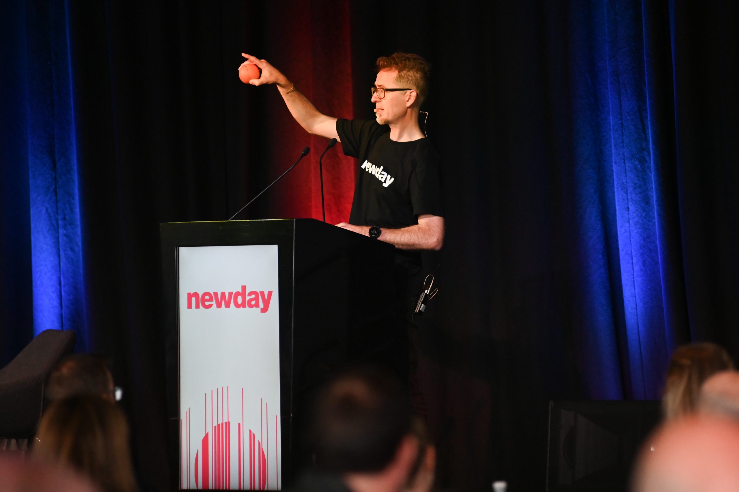 Man behind a podium holding orange sphere and gesturing to the sky