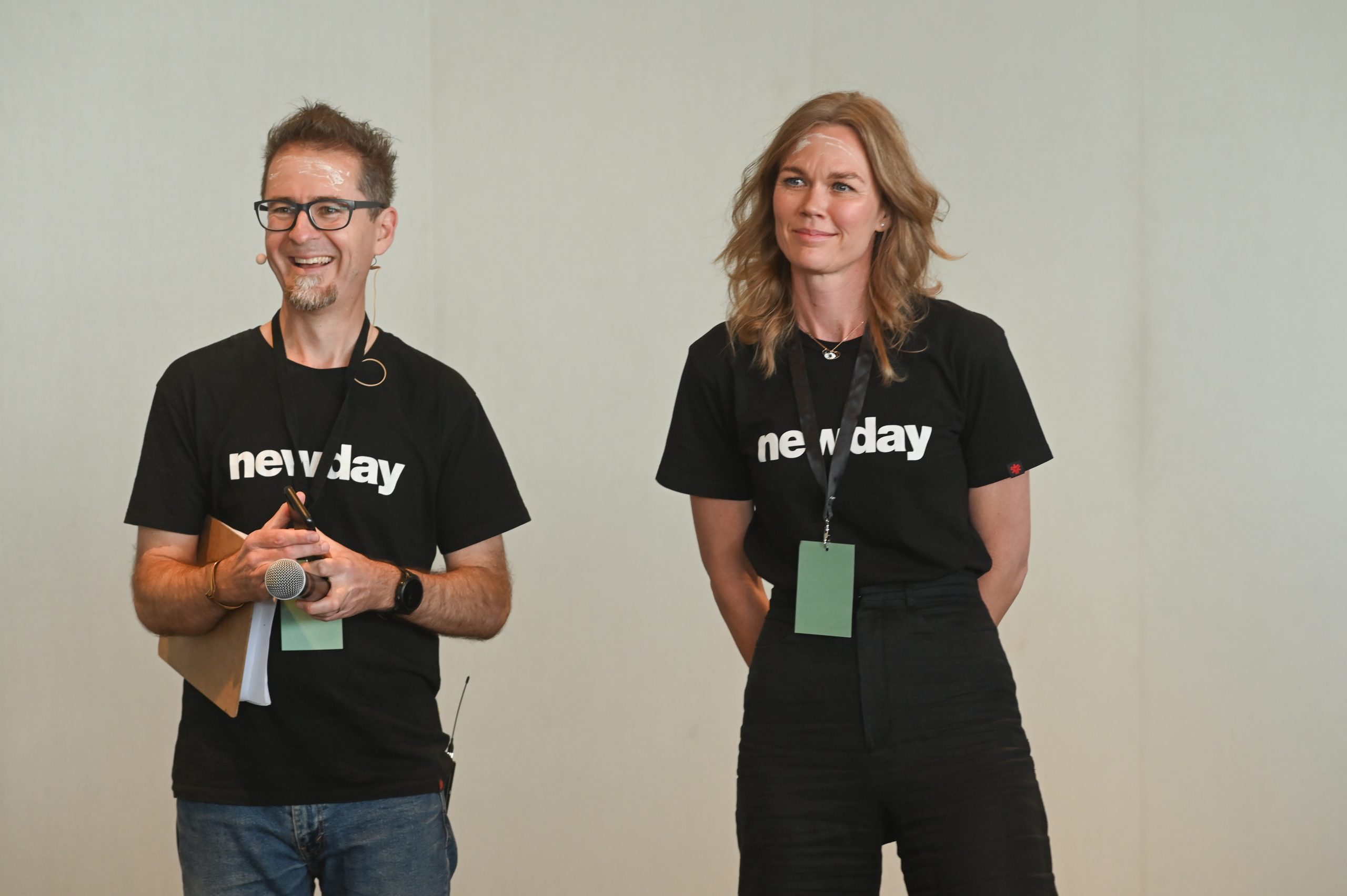 Two people wearing black t-shirts emblazoned with the word 'Newday' smile at someone off-camera. Each has pale ochre markings on their foreheads.